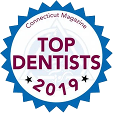 The image displays a blue ribbon with text on it stating  CONNECTICUT MAGAZINE TOP DENTISTS 2019  at the top, followed by the word  TOP  in large white letters and  DENTISTS  below it in smaller white letters. Below this title, there is a circular emblem with a blue border featuring a white star on the left side and a red seal on the right. Inside the emblem, there are two lines of text  the upper line reads  CONNECTICUT MAGAZINE  in bold white letters, and the lower line reads  TOP DENTISTS 2019  in smaller white letters. The background of the image is white with a faint pattern. The style of the image is graphic and promotional.