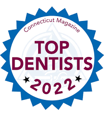The image shows a badge or emblem with text that reads 'Top Dentists 2022' at the top, followed by 'Connecticut Magazine' and below that, the words 'Dental Professionals.' The design includes a blue seal with stars around the perimeter, and there is a logo resembling a shield in the center. The badge has a red ribbon-like border with text on it indicating 'Top Dentists 2022' again. The background of the image is white.