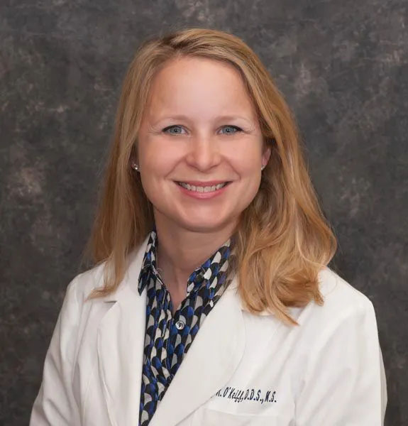 The image shows a woman wearing a white lab coat with name tags, standing against a background with a dark color scheme.