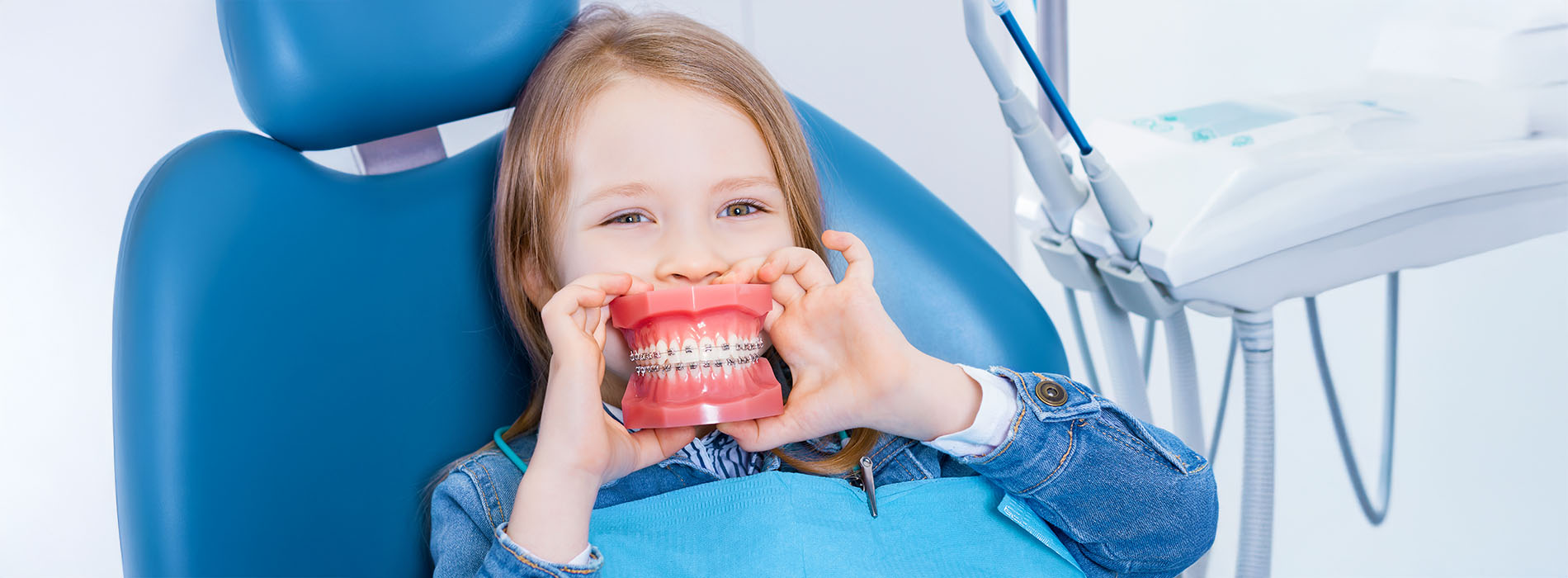 A young girl sitting in a dental chair with her mouth open, holding a toothbrush, surrounded by dental equipment.