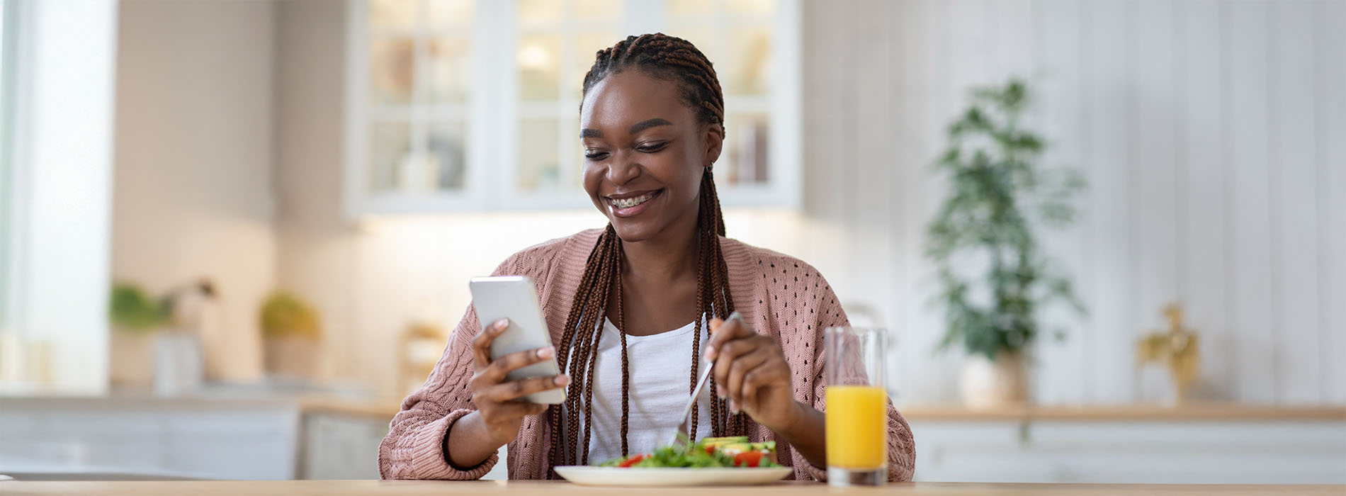 A woman sits at a kitchen counter, holding a glass of orange juice, with a plate of food before her, smiling towards the camera.