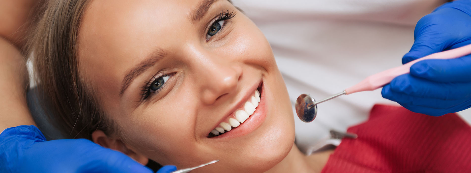 A woman with blonde hair smiling at the camera while sitting in a dental chair with a dentist s tool in her mouth.