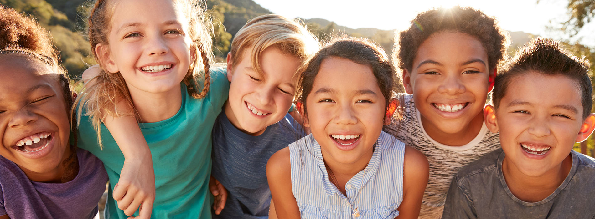 The image features four children smiling at the camera against a sunny background with a clear sky.