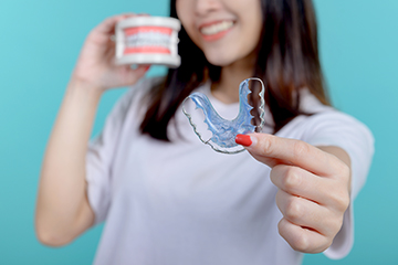 This is an image of a person holding a transparent object resembling a dental retainer with their right hand against a blue background, smiling at the camera.