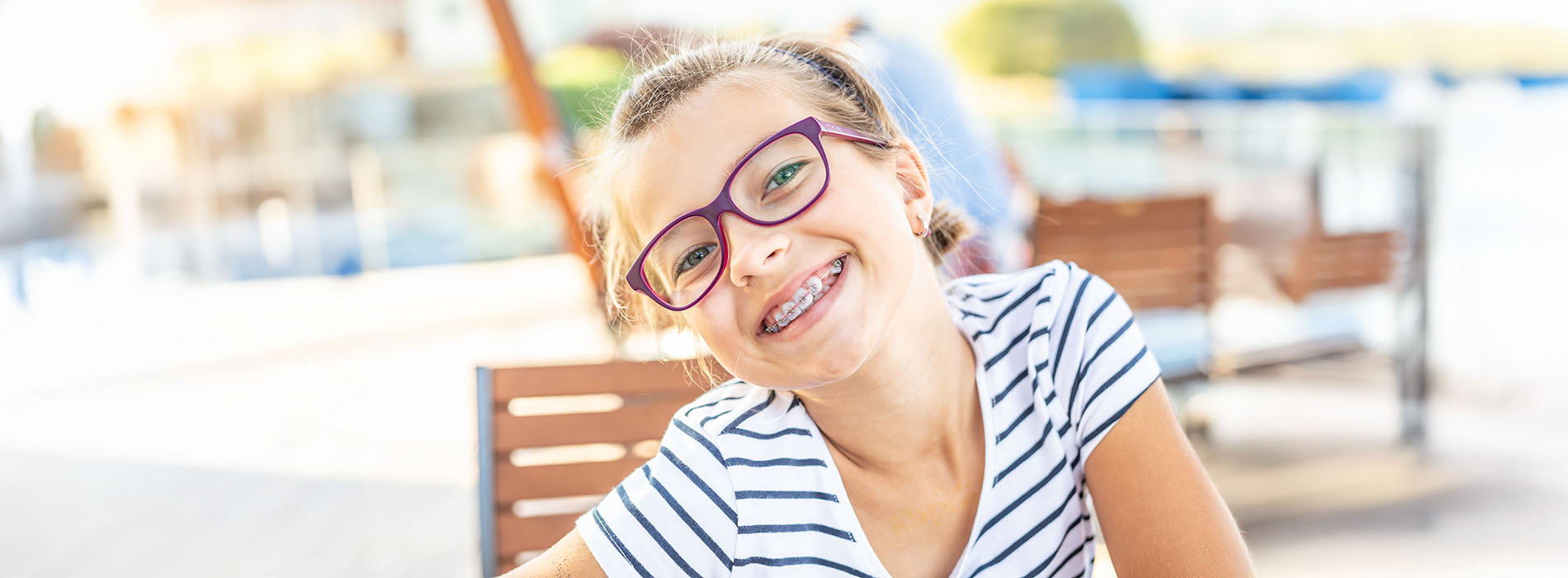 A young girl with glasses smiling at the camera while sitting outside on a bench.