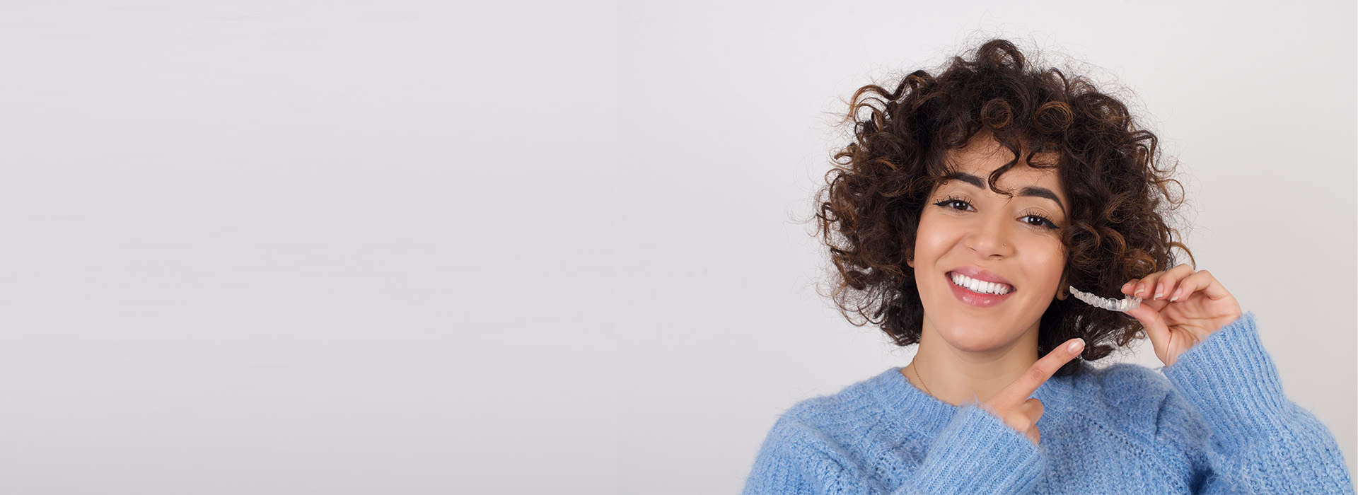 The image shows a smiling individual with dark hair, wearing a blue sweater, against a plain background.