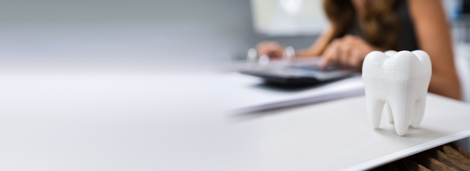 The image shows a person sitting at a desk working on a laptop with a whiteboard in the background, and there s a small figurine of a tooth near the top right corner.