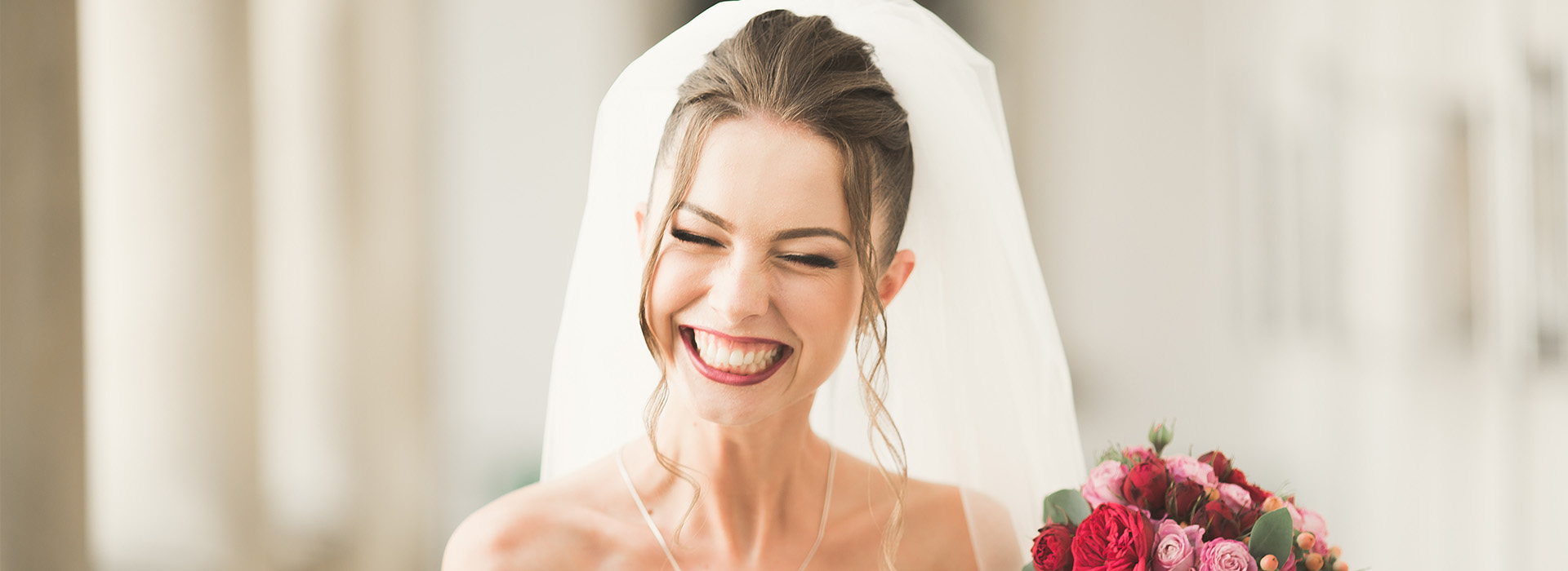 A bride wearing a white veil and laughing while holding flowers.