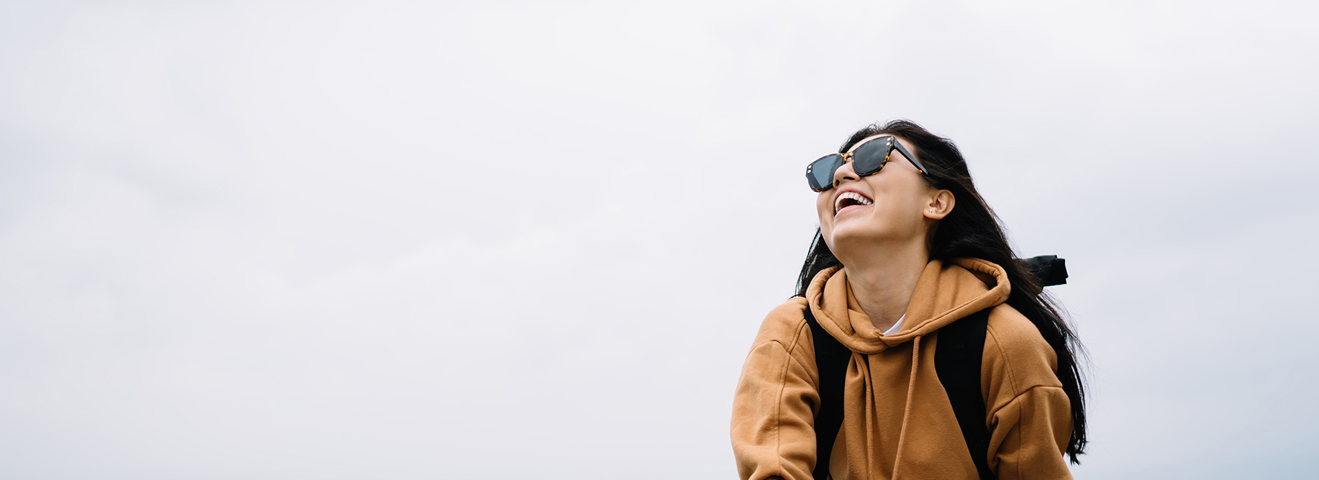 A person wearing sunglasses is standing against a cloudy sky with their back to the camera, smiling at something off-frame.