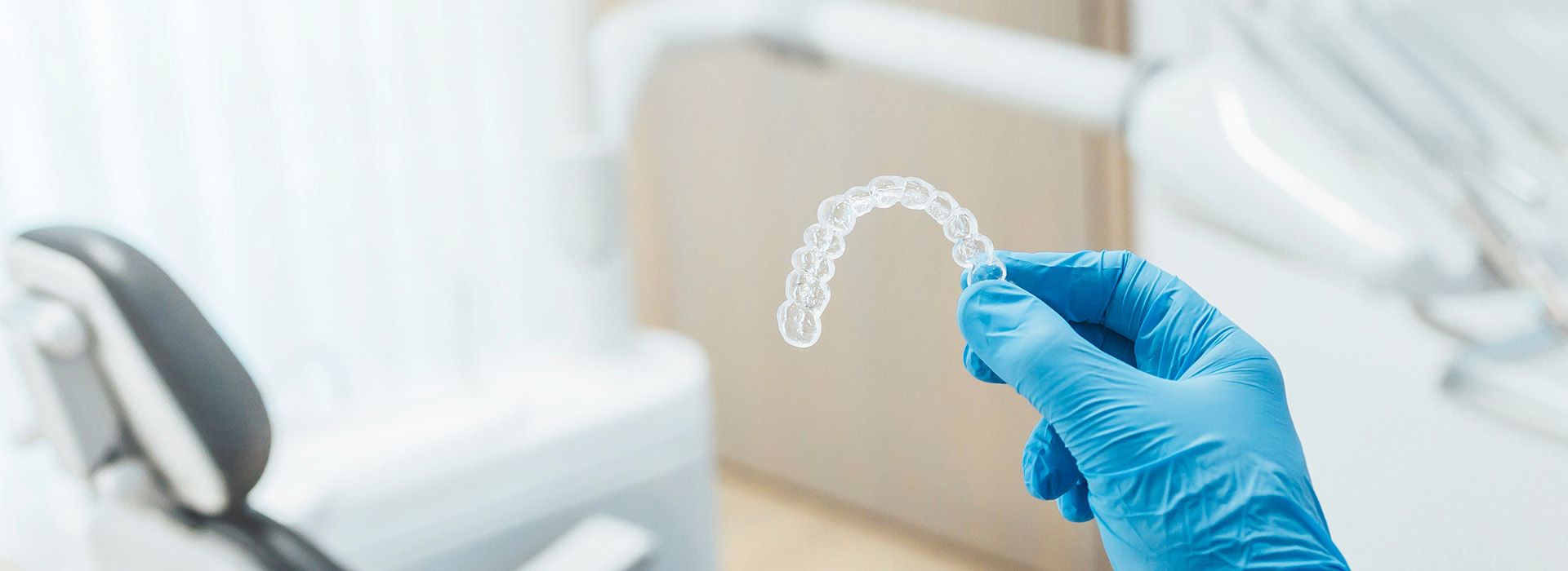 A person s hand holding a transparent object, likely a dental impression, against a blurred background with medical equipment.