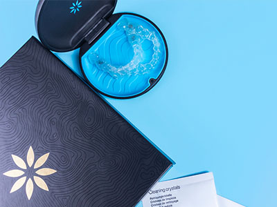 An open blue toothbrush with a clear bristle head next to a black box with a white logo on a teal background, all set against a light blue surface.