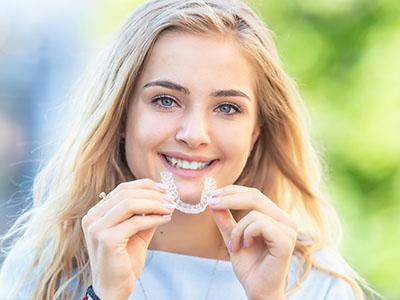 A young woman with blonde hair smiling at the camera while holding a dental retainer.