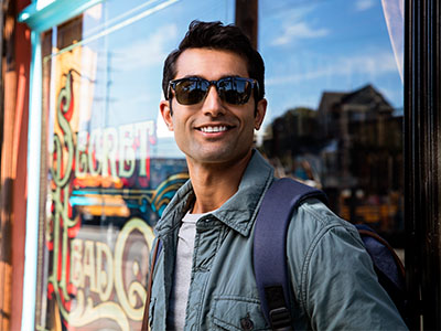 The image shows a man standing outside a storefront, wearing sunglasses, a backpack, and a light blue jacket, with a confident expression on his face.