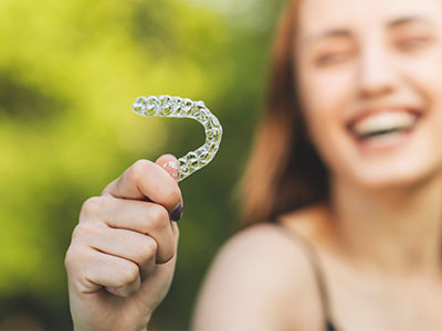 The image shows a smiling person holding up a clear plastic dental retainer with their left hand against a blurred outdoor background.