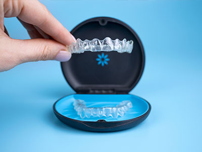 A hand holding a clear plastic tray with a blue toothbrush, a black plastic container with a white dental retainer inside, and a blue circular object resembling a mouthguard.