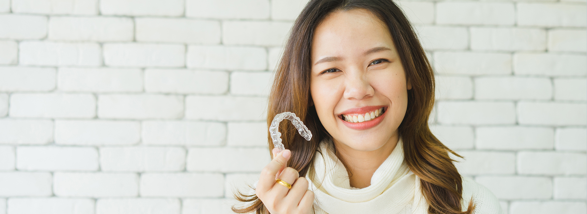 A woman wearing a white top smiles while holding a ring, with a blurred background featuring a brick wall and a white surface.