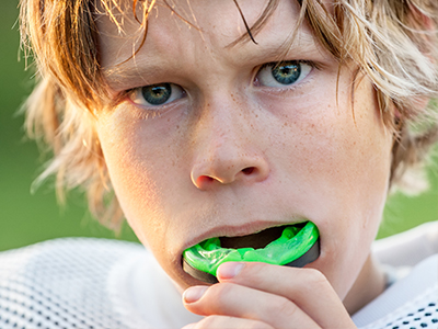 A young boy with blonde hair and blue eyes, wearing a football uniform, holding a toothbrush in his mouth while looking directly at the camera.