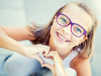 A young girl wearing glasses, smiling, and making a heart gesture with her hands.